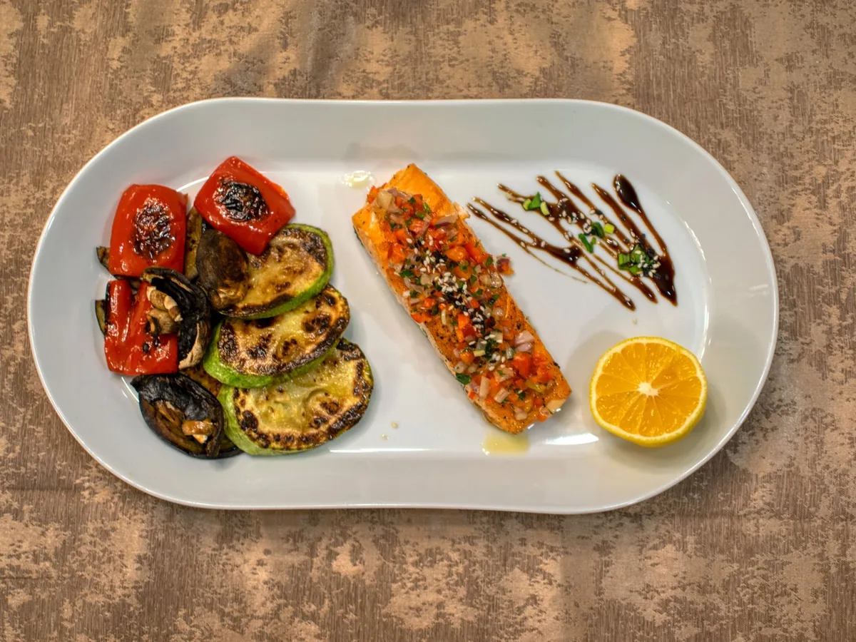 Gourmet grilled salmon fillet served with roasted vegetables and a lemon slice on a white plate.