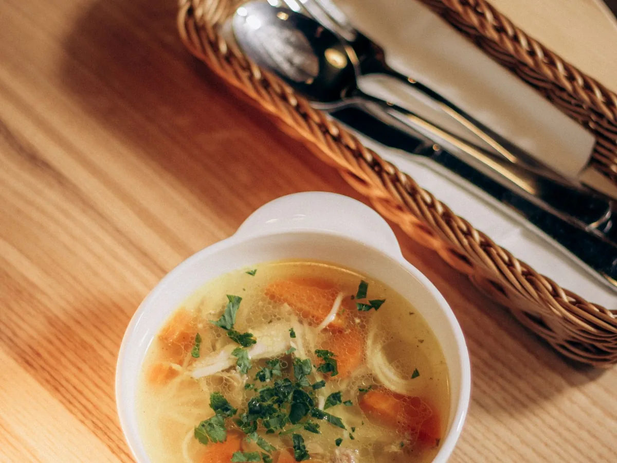 A warm bowl of chicken noodle soup with carrots and herbs on a wooden table