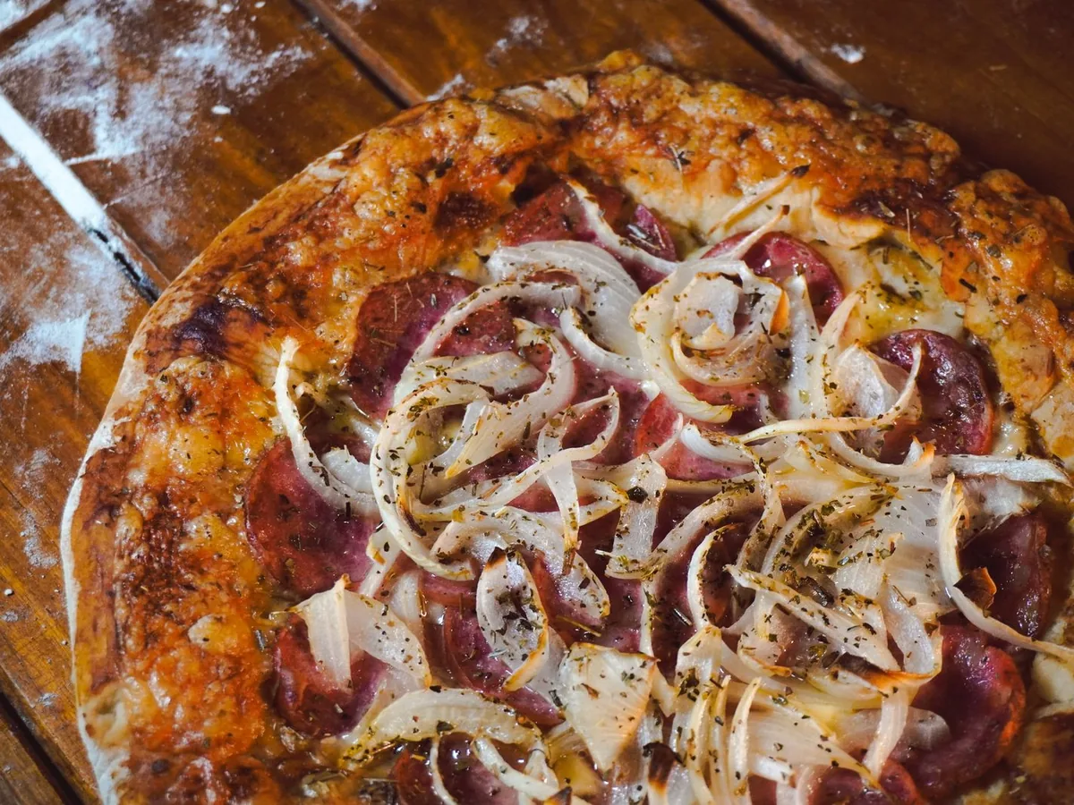 A homemade pepperoni pizza topped with onions and herbs on a rustic wooden table