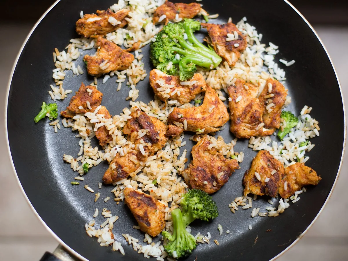 A black frying pan filled with white rice, chicken pieces and broccoli florets