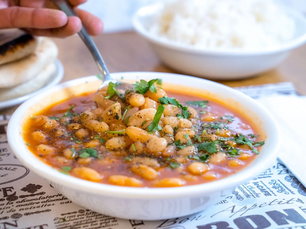 White bowl of white bean stew in a tomato broth garnished with fresh herbs, served with flatbread and rice.