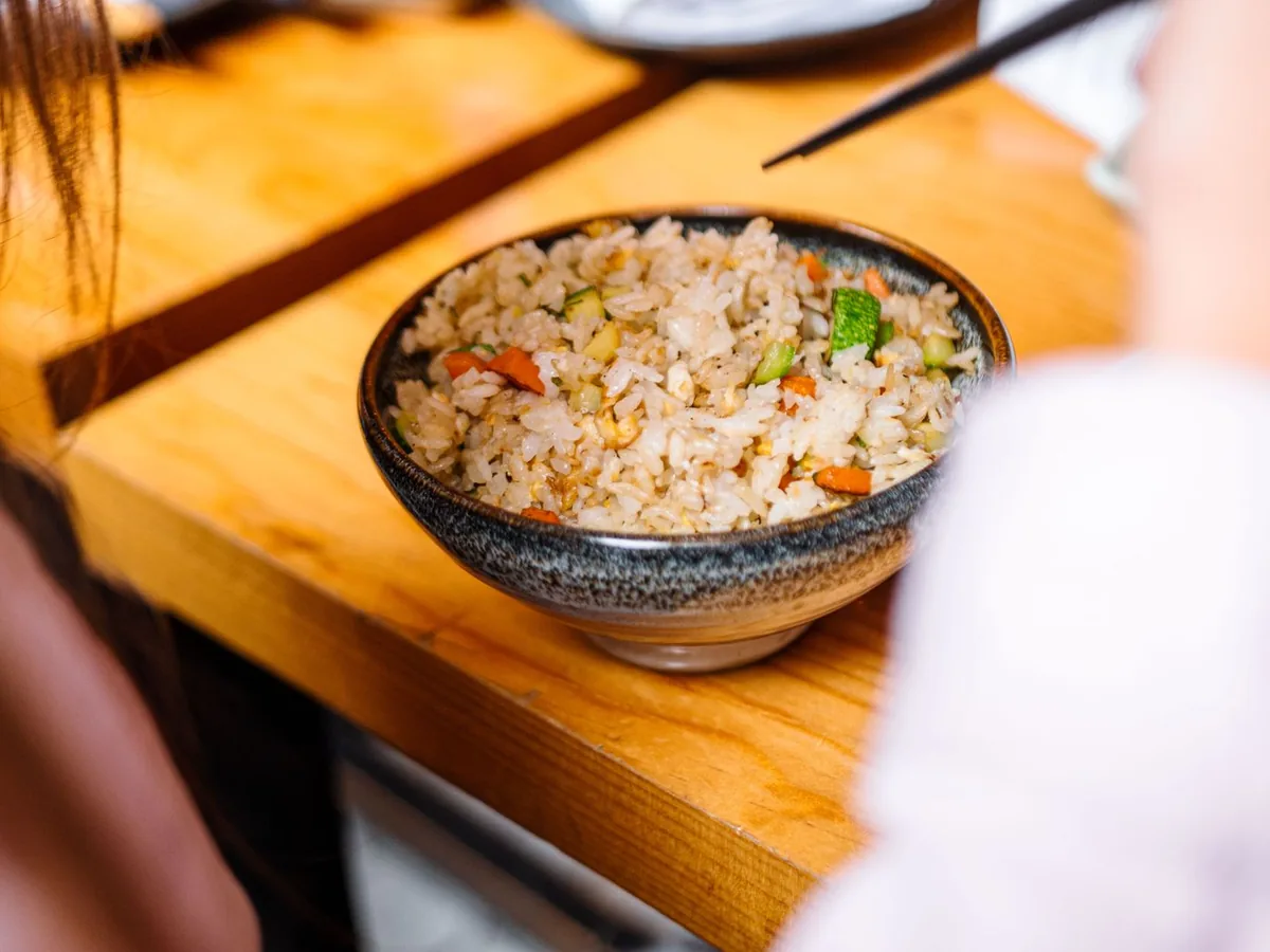 A close-up of a bowl of vegetable fried rice with carrots, zucchini and peas on a wooden table