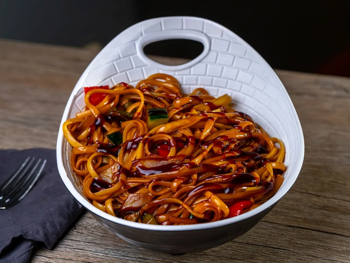 A bowl of stir-fried noodles with vegetables on a wooden surface